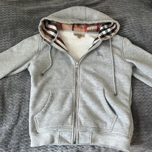 Grå hoodie från Burberry med dragkedja - Säljer en grå hoodie från Burberry med klassiskt rutigt foder i huvan och dragkedja framtill. Tröjan har två fickor, justerbar huva med snören och ribbade muddar. Perfekt för en stilren och bekväm look.