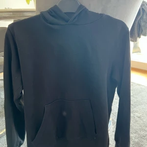 Svart hoodie med magficka - En klassisk svart hoodie med lång ärm och stor huva. Tröjan har en rymlig magficka framtill och är tillverkad i mjukt material som passar perfekt för vardagsbruk eller träning.