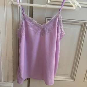 Ljuslila linnetopp med spets från Vero Moda - Säljer en ljuslila linnetopp från Vero Moda med tunna axelband och vacker spetsdetalj längs urringningen. Toppen är ärmlös och har en mjuk, slät yta som känns skön mot huden. Perfekt för varma dagar eller som lager under en kofta. Storlek S💗