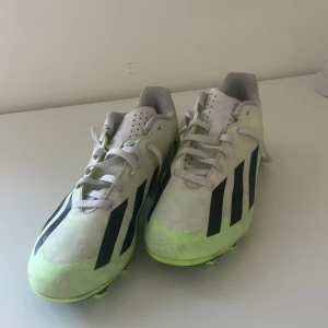 Vita och neongula fotbollsskor från Adidas - Säljer ett par vita Adidas fotbollsskor med svarta ränder och neongula detaljer. Skorna har snörning och är designade för bra grepp på planen. Perfekta för dig som vill sticka ut med stil på fotbollsplanen.
