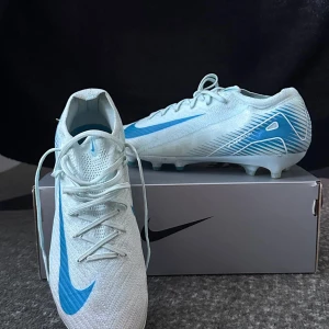 Nike Mercurial Elite - Säljer ett par Nike Mercurial fotbollsskor i vitt med blå detaljer och logga. Skorna har snörning och är designade för optimal prestanda på planen med en lätt ovandel och greppig sula. Perfekta för dig som vill ha stil och funktion på fotbollsplanen.