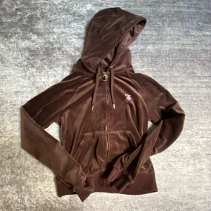 Brun velour hoodie från Juicy Couture - Mörkbrun hoodie i mjuk velour från Juicy Couture med glittrande Juicy-tryck på ryggen och JC-detaljer framtill. Dragkedja, huva och fickor. Perfekt för en avslappnad och trendig look. Lägger ut för 700 pga av den knappt är använd💗