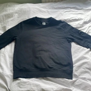 Svart sweatshirt från C.P. Company - Säljer en svart sweatshirt från C.P. Company i storlek M. Tröjan har rund halsringning, ribbade muddar och en ikonisk linsdetalj på ena ärmen. Perfekt för en stilren och avslappnad look. Självklart äkta och finns verifiering genom qr kod. Kom privat vid frågor eller funderingar!😊