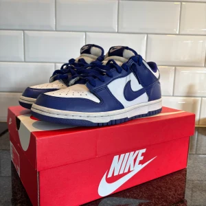 Nike Dunk Low blå och vita sneakers - Säljer ett par Nike Dunk Low sneakers i blått och vitt. Skorna har klassisk låg profil, vit bas med blåa overlays och matchande blå snörning. Perfekta för dig som gillar streetwear och vill ha en ikonisk look. Kom med bud !!!!!