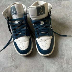 Nike Air Jordan 1 Mid blå och vita sneakers - Säljer ett par Nike Air Jordan 1 Mid sneakers i blått och vitt. Skorna har högt skaft, klassisk snörning och den ikoniska swoosh-loggan på sidan. Perfekta för dig som gillar streetwear och vill ha en stilren look. Dessa är använda 3 gånger sedan endast stått hemma, så är i otroligt bra skick! Storlek 40.