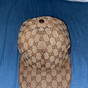 Beige keps från Gucci med GG-mönster - Säljer en beige keps från Gucci med klassiskt GG-monogram över hela kepsen och gröna/röda ränder på sidan. Kepsen är i bomullsmaterial och har en böjd skärm. Perfekt för dig som gillar lyxiga accessoarer.