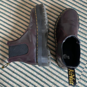 Vinröda Chelsea boots från Dr. Martens - Säljer ett par vinröda Chelsea boots från Dr. Martens med klassisk gul söm och grov sula. Skorna har elastiska paneler på sidorna och dragflik bak. Ovanligt bekväma för att vara Dr. Martens och väldigt sparsamt använda! 