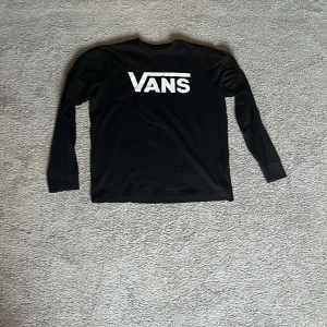 Svart långärmad tröja från Vans - Svart långärmad tröja från Vans med stor vit logga på bröstet. Klassisk passform och rund halsringning. Perfekt för en avslappnad streetwear-look.