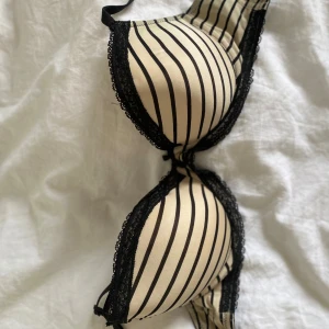 Randig bh med spetskant från H&M - Säljer en snygg bh från H&M i beige med svarta vertikala ränder och svarta spetsdetaljer längs kanterna. Bh:n har vadderade kupor och justerbara axelband. Perfekt för dig som gillar en klassisk men ändå trendig look.