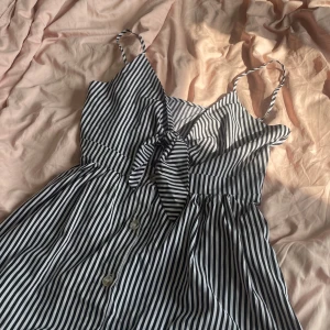 Randig klänning - Säljer en svartvit randig klänning med smala axelband och knyt framtill. Klänningen har knappar längs hela framsidan och en vid kjol som går ner till smalbenen. Perfekt för varma dagar och sommarfester!