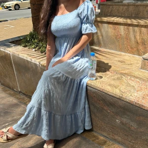 Ljusblå långklänning off shoulder  - Säljer en ljusblå långklänning med smock upptill och korta puffärmar. Klänningen har en av axlarna bar och är tillverkad i ett lätt, flowy material. Perfekt för varma dagar och har en romantisk vibe.