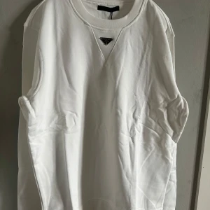 Vit crewneck hoodie med Prada logo. - Säljer en stilren vit sweatshirt från Prada med klassisk logga i metall framtill. Tröjan har rund halsringning och är tillverkad i mjuk bomull. Perfekt för en trendig och avslappnad look.