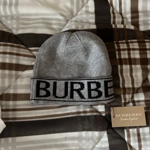 Grå mössa från Burberry - Snygg grå mössa från Burberry med bred uppvikt kant och stor svart logotyp runtom. Tillverkad i mjukt stickat material som känns skönt mot huden. Perfekt accessoar för kyliga dagar.