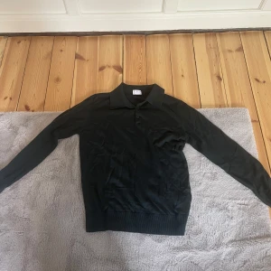 Svart pikétröja från Asket - Klassisk svart pikétröja från Asket i mjukt material. Tröjan har krage, knäppning framtill och långa ärmar. Perfekt för en stilren och enkel look.