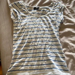 Vit och blå randig t-shirt från JC - Vit t-shirt med blå horisontella ränder från JC. T-shirten har korta ärmar och rund halsringning. Perfekt för en avslappnad stil under varmare dagar. OBS! en liten svart fläck