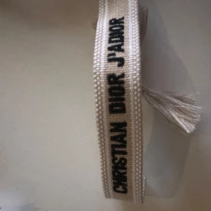 Beige armband från Christian Dior - Säljer ett beige armband från Christian Dior med texten 'J'ADIOR' i svart. Armbandet har tofsar i ändarna och är gjort i ett vävt material. Perfekt accessoar för att lyfta din outfit.