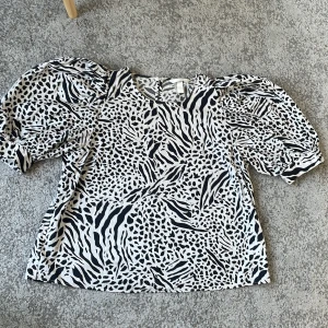 Svartvit blus med puffärmar från H&M - Säljer en svartvit blus från H&M med djurmönster och stora puffärmar. Blusen har rund halsringning och knäppning bak i nacken. Perfekt för dig som vill sticka ut med ett coolt mönster.
