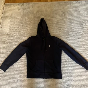 Svart hoodie från Polo Ralph Lauren - Säljer en svart hoodie från Polo Ralph Lauren med dragkedja och huva. Tröjan har fickor framtill och det klassiska broderade logotypen på bröstet. Finns både med lång och kort ärm, perfekt för olika stilar.