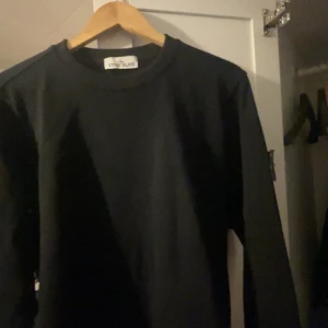 Svart sweatshirt från Stone Island - Säljer en svart sweatshirt från Stone Island med klassisk rund halsringning och lång ärm. Tröjan har en enkel design med logotyp-patch på ena ärmen. Perfekt för en stilren och avslappnad look.