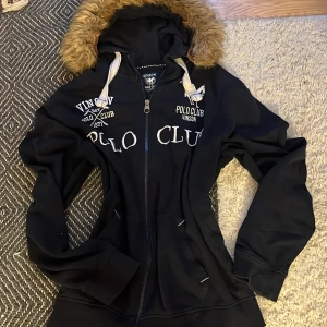 Svart hoodiejacka med fuskpäls från Vinson Polo Club - Svart hoodiejacka från Vinson Polo Club med dragkedja och stor huva kantad med brun fuskpäls. Jackan har broderade loggor och text på bröstet samt vita snören vid huvan. Perfekt för kyliga dagar.