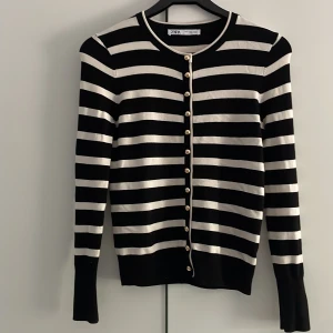 Svartvit randig kofta från Zara - Säljer en svart och vit randig kofta från Zara med guldknappar framtill. Koftan har långa ärmar och rund halsringning. Perfekt att styla med jeans eller kjol för en snygg look.