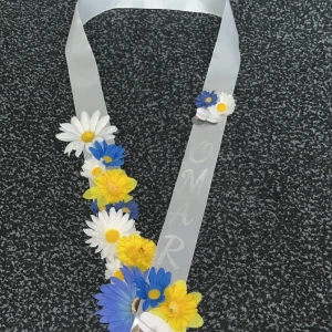 Studentband med blommor i blått, vitt och gult - Studentband i vitt med konstgjorda blommor i blått, vitt och gult. Bandet har texten 'OMAR' och är dekorerat med prästkragar och andra färgglada blommor, perfekt till studenten.