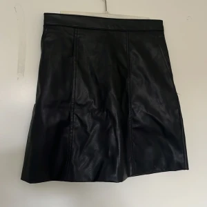 Svart skinnkjol från H&M Divided - Säljer en svart skinnkjol från H&M Divided. Kjolen är kort med en rak passform och har en synlig dragkedja bak. finns även 1 ficka på båda sidorna. aldrig använd