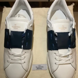 Valentino Garavani Open sneakers med blå detalj - Säljer ett par vita Valentino Garavani Open sneakers med bred blå läderdetalj över snörningen. Skorna har rund tå, platt sula och klassisk logotyp på plösen. Perfekta för dig som gillar stilrena och exklusiva sneakers.