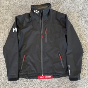 Svart seglarjacka från Helly Hansen - Svart vindjacka från Helly Hansen med röda dragkedjor och logga på kragen, ärmen och nederkanten. Jackan har hög krage, tre fickor med dragkedja och meshfoder på insidan. Perfekt för blåsiga dagar. Jackan har inga synliga defekter mer än lite smuts! Hör av dig vid minsta fråga! 