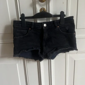 Lågmidjade Svarta jeansshorts  - Snygga svarta Lågmidjade jeansshorts med råa fransade kanter. Perfekta för varma dagar och passar till det mesta. Stängs med knapp och dragkedja framtill. Säljer då dem inte passar. Bra skick , använda fåtal gånger.