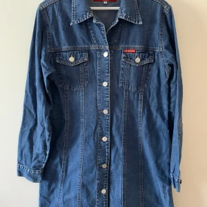 Jeansklänning från B. DENIM by B.YOUNG - Säljer en klassisk jeansklänning från B. DENIM by B.YOUNG i mellanblå denim. Klänningen har långa ärmar, knappar hela vägen framtill och två bröstfickor med lock. Perfekt för en avslappnad och cool look.