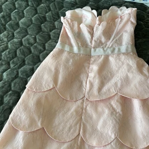 Ljusrosa klänning med spets och rosett från ASOS Luxe - Supersöt ljusrosa klänning från ASOS Luxe med spetsdetaljer, volangliknande vågade kanter och en söt rosett i midjan. Axelbandslös modell med markerad byst och kort längd. Perfekt för speciella tillfällen! Köpt för 700kr och haft på mig en gång 