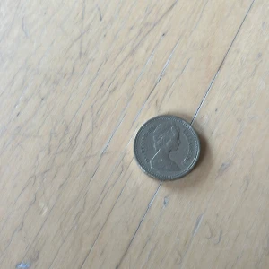 Brittiskt mynt - One Pound 1983 - Säljer ett brittiskt enpundsmynt i metall med drottningens porträtt på ena sidan och ett kungligt vapen på andra sidan. Myntet är runt och har text ingraverad på kanten. Perfekt för samlare eller som souvenir.