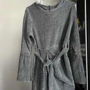 Glittrig silverklänning med knytband - Säljer en glittrig silverfärgad klänning med lång ärm och knytband i midjan. Klänningen har en rund halsringning och vida ärmslut som ger en trendig look. Perfekt för dig som vill sticka ut med stil.