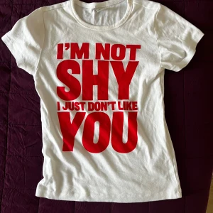 Vit t-shirt med röd text - Vit t-shirt med stor röd text på framsidan: 'I'M NOT SHY I JUST DON'T LIKE YOU'. T-shirten har en klassisk rund hals och korta ärmar. Perfekt för dig som vill sticka ut med ett tydligt statement.
