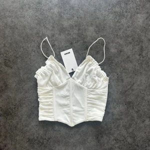 Vit croppad korsettopp - Säljer en vit croppad korsettopp med smala axelband och rynkade detaljer på sidorna. Toppen har en v-ringad design och är perfekt för att skapa en trendig look.