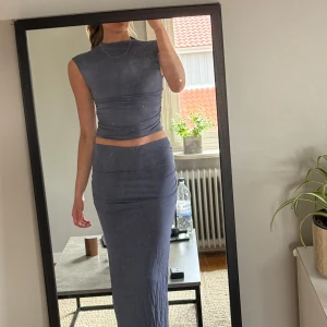 Set från Gina!  - Säljer en snygg tvådelad outfit i mörkgrått. Setet består av en ärmlös, tight cropptopp och en matchande långkjol med låg midja. Topp i M, kjol i S