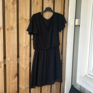 Svart kortklänning med volangärmar - Svart klänning med kort längd och volangärmar. Klänningen har en lös passform upptill och resår i midjan för en bekväm siluett. Perfekt för dig som gillar enkel men stilren design.