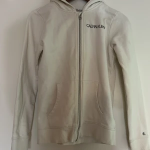 Vit hoodie från Calvin Klein - Säljer en stilren vit hoodie från Calvin Klein med dragkedja framtill och fickor. Märkets logga syns på bröstet och en patch med 'JEANS' på ryggen. Perfekt hoodie nu till sommarkvällarna🙌🏻🔥