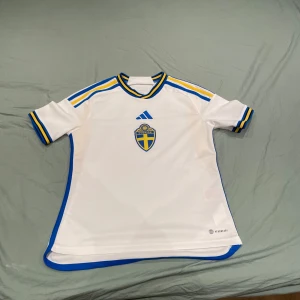 Svensk landslagströja i vitt med blå och gula detaljer - Vit fotbollströja från Adidas med Sveriges landslagsemblem på bröstet. Blå och gula ränder på axlar och ärmslut, samt blå kant längs sidorna. Klassisk landslagsdesign med texten 'Sverige' i nacken.