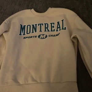 Vit sweatshirt med tryck från Gina Tricot - Säljer en vit sweatshirt från Gina Tricot i storlek S. Tröjan har tryck med texten 'Montreal Sports Champ' i grönt framtill. Klassisk rund halsringning och långa ärmar. Perfekt för en avslappnad stil.
