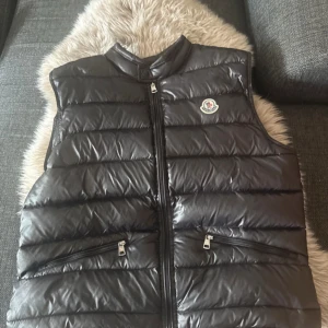 Svart dunväst från Moncler - Snygg svart dunväst från Moncler med ståkrage och dragkedja framtill. Västen har två fickor med dragkedja och klassisk Moncler-logga på bröstet. Perfekt för lager-på-lager under kyliga dagar.