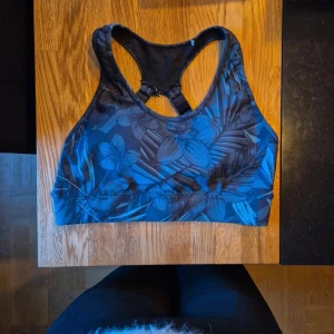 Blå sport-bh med blommigt mönster - Sportbh från OTP i fint skick. Mörkblå med bladigt mönster.                          Bh:n har brottarrygg och justerbara axelband för extra stöd. Materialet är stretchigt och består av 80% polyester och 20% spandex.    Den är hel Storlek S 36/38 går det att utläsa även fast det va svårt att få på bild.