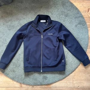 Les dexus tröja  - Säljer en stilren mörkblå track jacket från Les Deux med dragkedja framtill och ståkrage. Perfekt nu till sommaren 