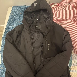 Svart jacka från Sail Racing - Svart jacka från Sail Racing med huva och dragkedja framtill. Jackan har en sportig look och logga på bröstet samt huvan. Perfekt för kyliga dagar.