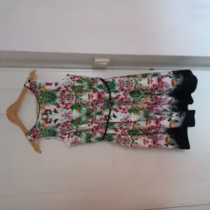 Blommig klänning i bomull/elastan med utställd kjol. Tyget har lite stretch i sig. Dragkedja i ryggen och skärp i midjan. Tryck med blommor, löv och papegojor i rosa och grönt. 