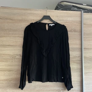 Blus  - Svart blus med volang ifrån axel till axel. Andra bilden är uppljust därför en konstig färg.