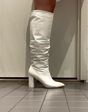 Vita knähöga stövlar - Vita stövlar i fuskläder, storlek 39. Klackhöjd 9cm. Endast använd ett fåtal tillfällen.  Färg bortskavd på tån, se sista bild! Annars i väldigt bra skick. Skickas mot frakt