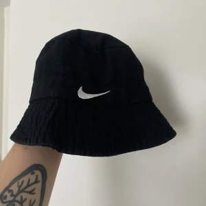Nike Buckethat - En svart buckethat/fiskarhatt med Nike logga bak och fram. Köpt secondhand, säljes då den är för liten. Har inga exakta mått men uppskattar att inreomfåget är drygt 48cm.  OBS Se bild 2, något tyg på insidan har lossnat lite. 
