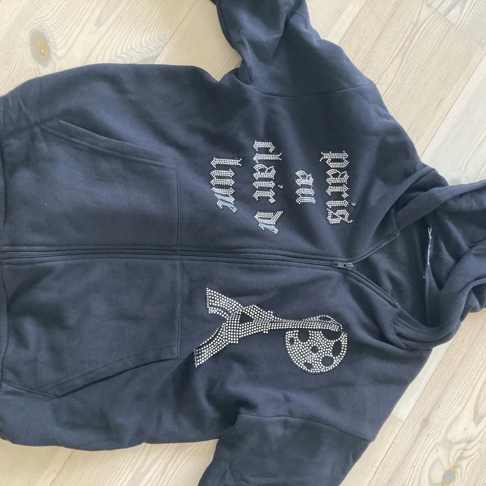 Oanvänd ny svart luvtröja med zip. Rhinestone i mönster. Storlek XS men mer lik en M. Inköpt för 699 kr.. Hoodies.